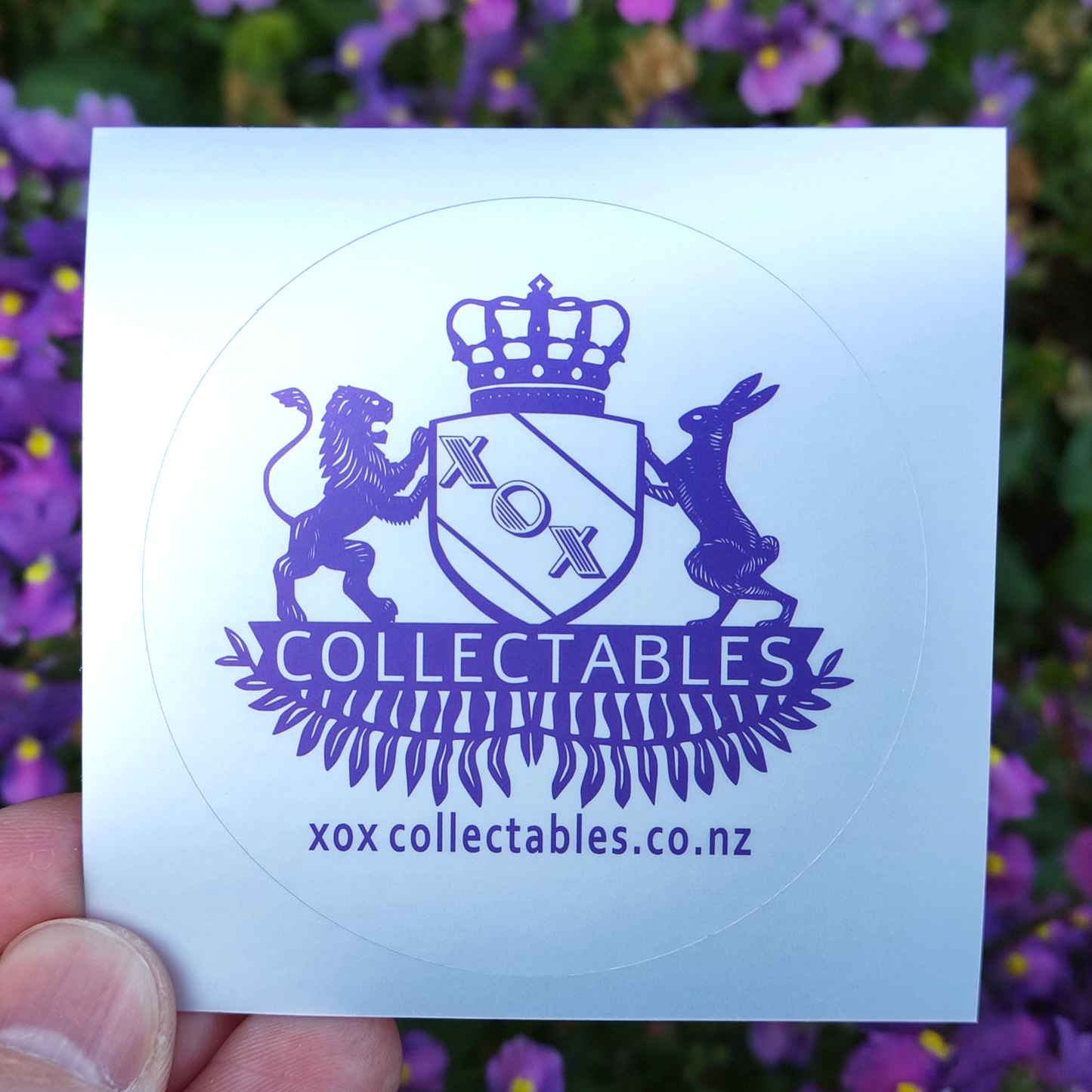 XOX Collectables Coat of Arms Sticker. Purple on reflective matte silver. In front of purple flowers