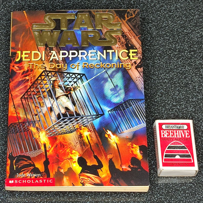 Book titled 'Jedi Apprentice: The Day of Reckoning' by3 John Jackson Miller on a dark textured surface with a deck of cards.
